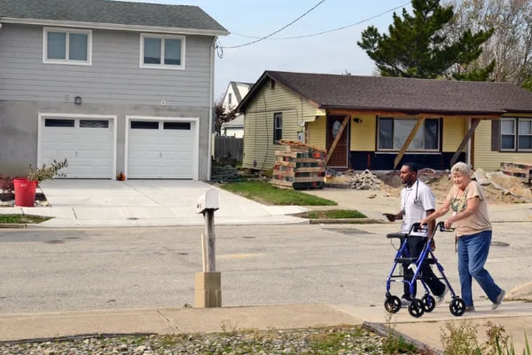 Barbara Fox walks along Cummings Place in Brigantine with her physical therapist Marvin Royal on October 29, 2014, on the second anniversary of Superstorm Sandy. Her home (not shown) is similar to the house on the right. Both will be raised, just like the home on the left. ( TOM GRALISH / Staff Photographer )