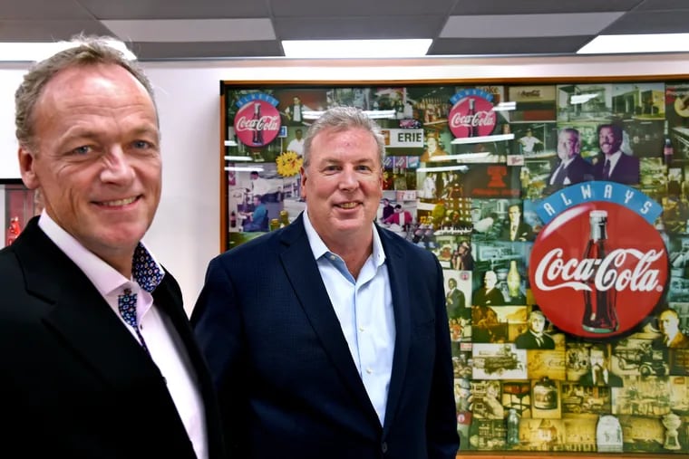 Paul Mulligan (left) and Fran McGorry (right) co-owners of Liberty Coca-Cola Beverages pause by a Coke mural in the lobby of their bottling plant.