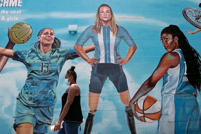 A pedestrian walks past a portion of a mural honoring women in sports and featuring U.S. Olympians, including  paralympian Oksana Masters (center). The mural is at the corner of Germantown Avenue and Price Street in Philadelphia. Left on the mural is soccer star Alex Morgan. Right is WNBA star Chiney Ogwumike.