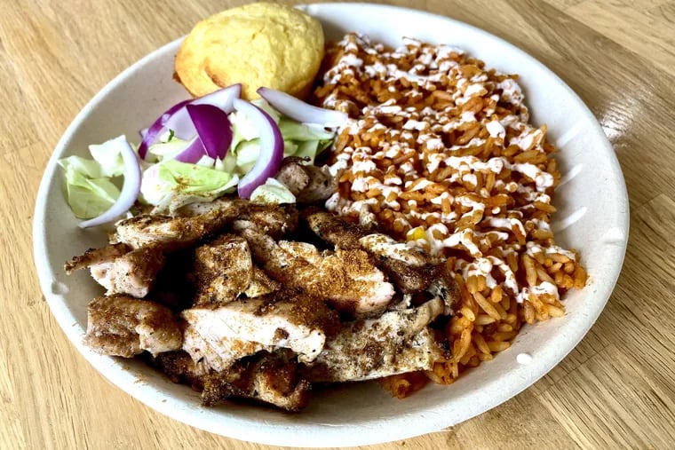 A chicken bowl at Suya Suya with jollof rice topped with white sauce, a side salad, and cornbread muffin.