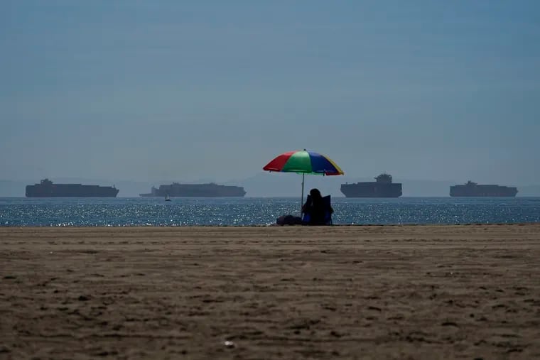 A beach-goer sits on the sand in Seal Beach Calif., as container ships waiting to dock at the Ports of Los Angeles and Long Beach are seen in the distance.