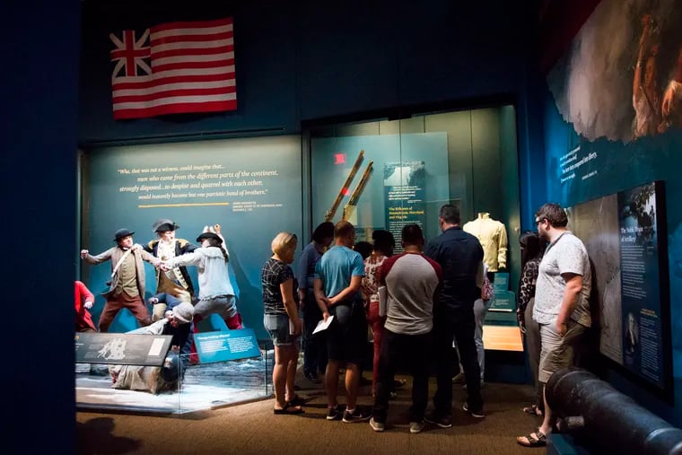 Joella Clamen, an educator at the Museum of the American Revolution, is crowded by participants as she explains the history of the U.S. flag. The museum is using its exhibits, artifacts, and stories to help prepare green-card holders for their naturalization tests.