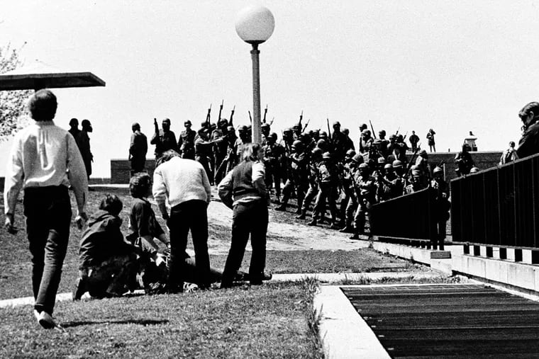 A group of youths cluster around a wounded person as Ohio National Guardsmen, wearing gas masks, hold their weapons in background on Kent State University campus in Kent, Ohio, Monday, May 4, 1970.  Members of the Guards killed four students and injured nine during a campus protest against the Vietnam War.  This picture was made by Kent State student photographer Douglas Moore.