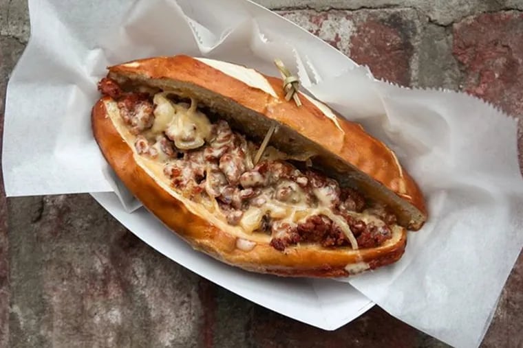 Bavarian cheesesteak on Frankford Hall's happy hour menu includes chopped bauernwurst with cheese sauce and onions on a pretzel roll. ⠀