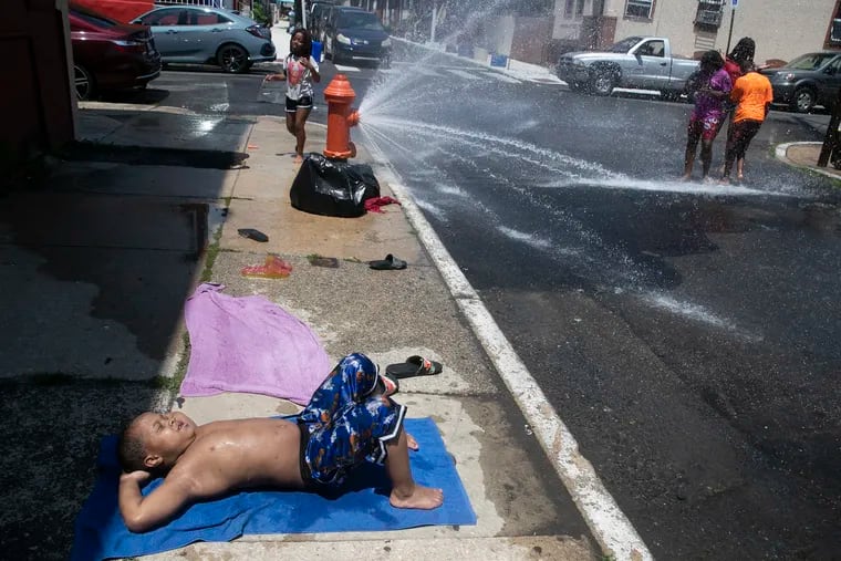 Zahir Garrett, 8, sunbathes on the street while kids play in an open fire hydrant in North Philadelphia. The Hunting Park neighborhood faces the highest heat index in the city, but will go another summer without its pool.