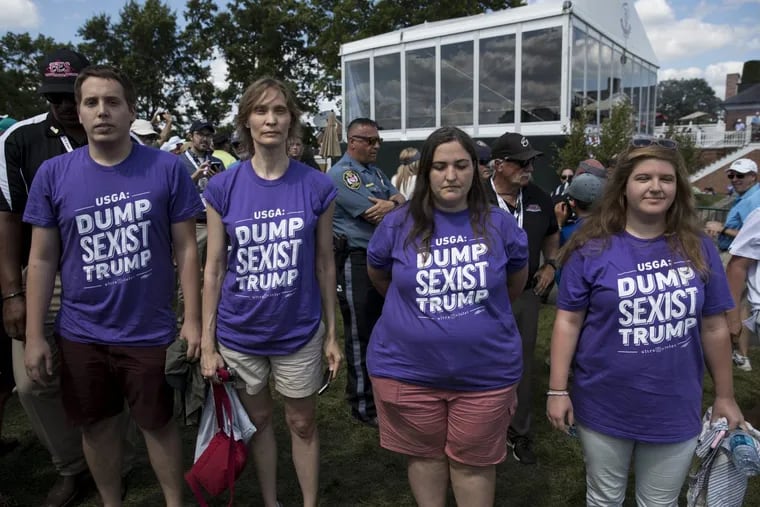 Protesters with t-shirts that read “USGA:Dump Sexist Trump” position themselves near the presidential viewing stand, behind, where President Donald Trump is watching the U.S. Women’s Open Golf at Trump National Golf Club.