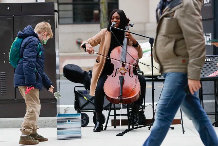 Cellist/vocalist Aijee Evans, watches Ben Maher, 9, drop dollar bills into a tip box, while Evans performed at Ninth and Market Streets in Center City on Monday, November 22, 2021.  Evans was robbed in November while performing at Fourth and South Streets by a couple of teens.
