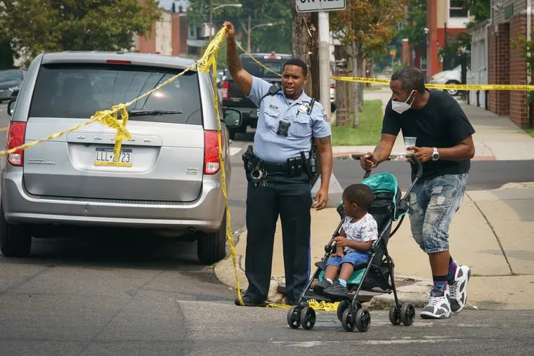 A police officer lifts crime scene tape for Ramon Harris and his son Sekani Brown-Harris, who he picked up from daycare at 56th and Vine Streets, on the same block where police were investigating a triple shooting, Wednesday, July 21, 2021.