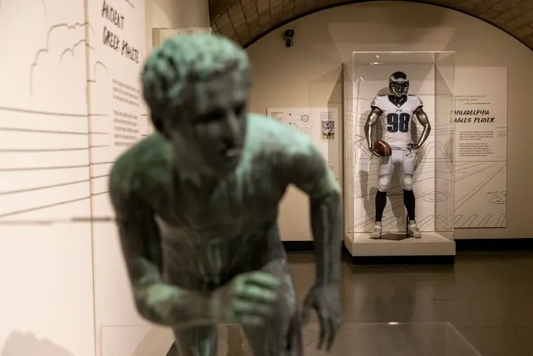 A statue of a naked ancient Greek athlete juxtaposed with athletes today, like players on the Eagles, with padding, cleats, helmets, jersey numbers, and other attire, at “The Stories We Wear” exhibition at the Penn Museum.