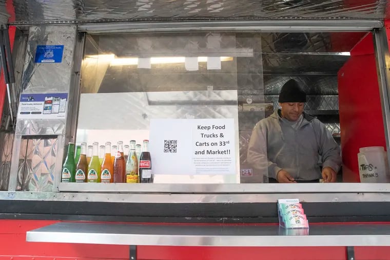 A sign urging to keep food trucks is seen on the Los Compadres cart where Alfonso Pantoja (right) is working outside of Drexel University at Market Street between 33rd and 34th in Philadelphia on Wednesday, Dec. 11, 2019. The food trucks there could be evicted if a City Council bill passes Thursday.