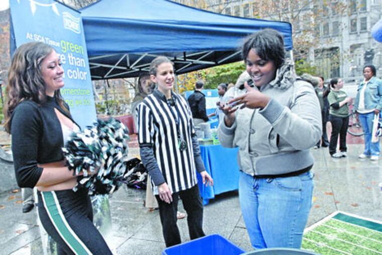 At a JFK Plaza recycling promotion, Eagles cheerleader Tina Thomas (left) and Raquel Carbonari watch as a Philadelphian named Sasha plays a game in which the goal is sorting recyclables.
