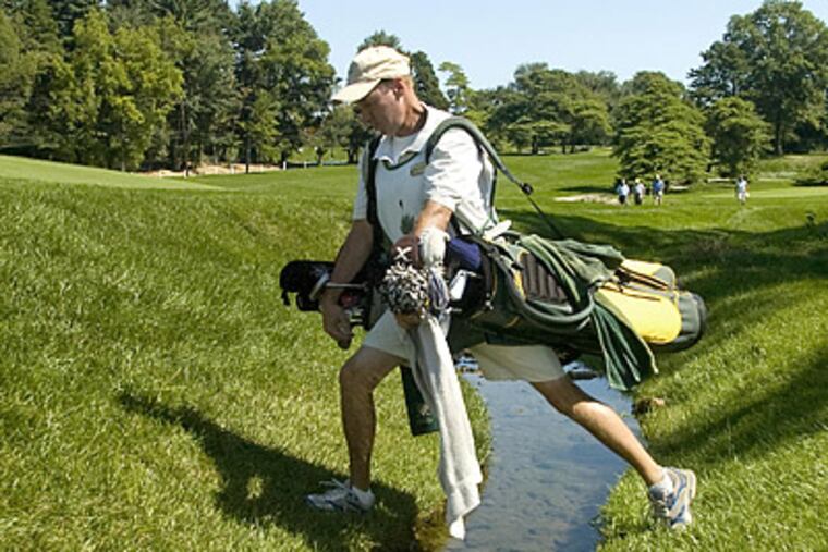 Merion caddy Jack Hopkins carries two bags across a stream on the fifth hole of the Merion East course. (Clem Murray/Staff Photographer)