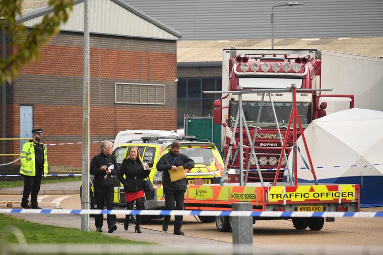 People walk past a police officer at the scene after a truck, seen in rear, was found to contain a large number of dead bodies, in Thurrock, South England, early Wednesday Oct. 23, 2019. Police in southeastern England said that 39 people were found dead Wednesday inside the truck container believed to have come from Bulgaria.
