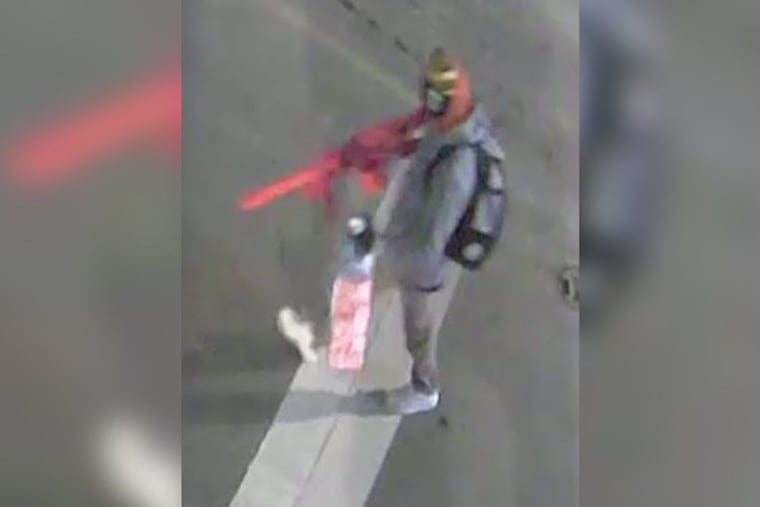 A screenshot of a surveillance video captures the suspect in a recent vandalism incident, where an unknown white male painted racist and antisemitic slogans on the exterior walls of Roxborough High School on Jan. 4, 2026. Police describe the suspect as a white male, wearing an orange scarf/wrap, green and black winter hat, gray hooded jacket, gray pants, and a gray and black backpack.