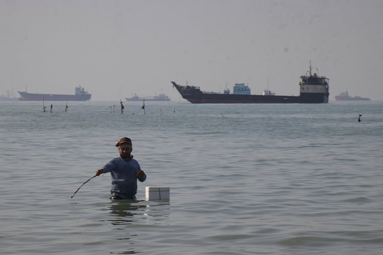 A man stands in the water, appearing to fish, as bulk carriers, cargo ships, and service vessels line the horizon in the Strait of Hormuz off Bandar Abbas, Iran, Monday, April 27, 2026.