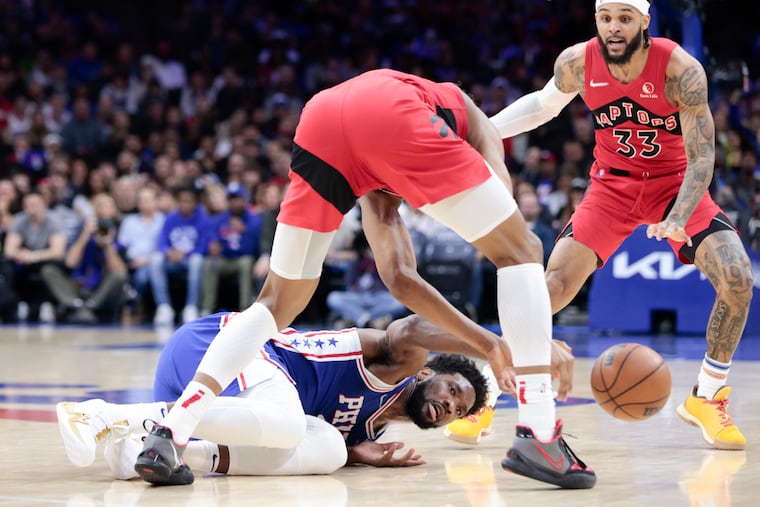 Sixers center Joel Embiid reaches for the loose basketball on the floor against Toronto Raptors forward Scottie Barnes and Toronto Raptors guard Gary Trent Jr.