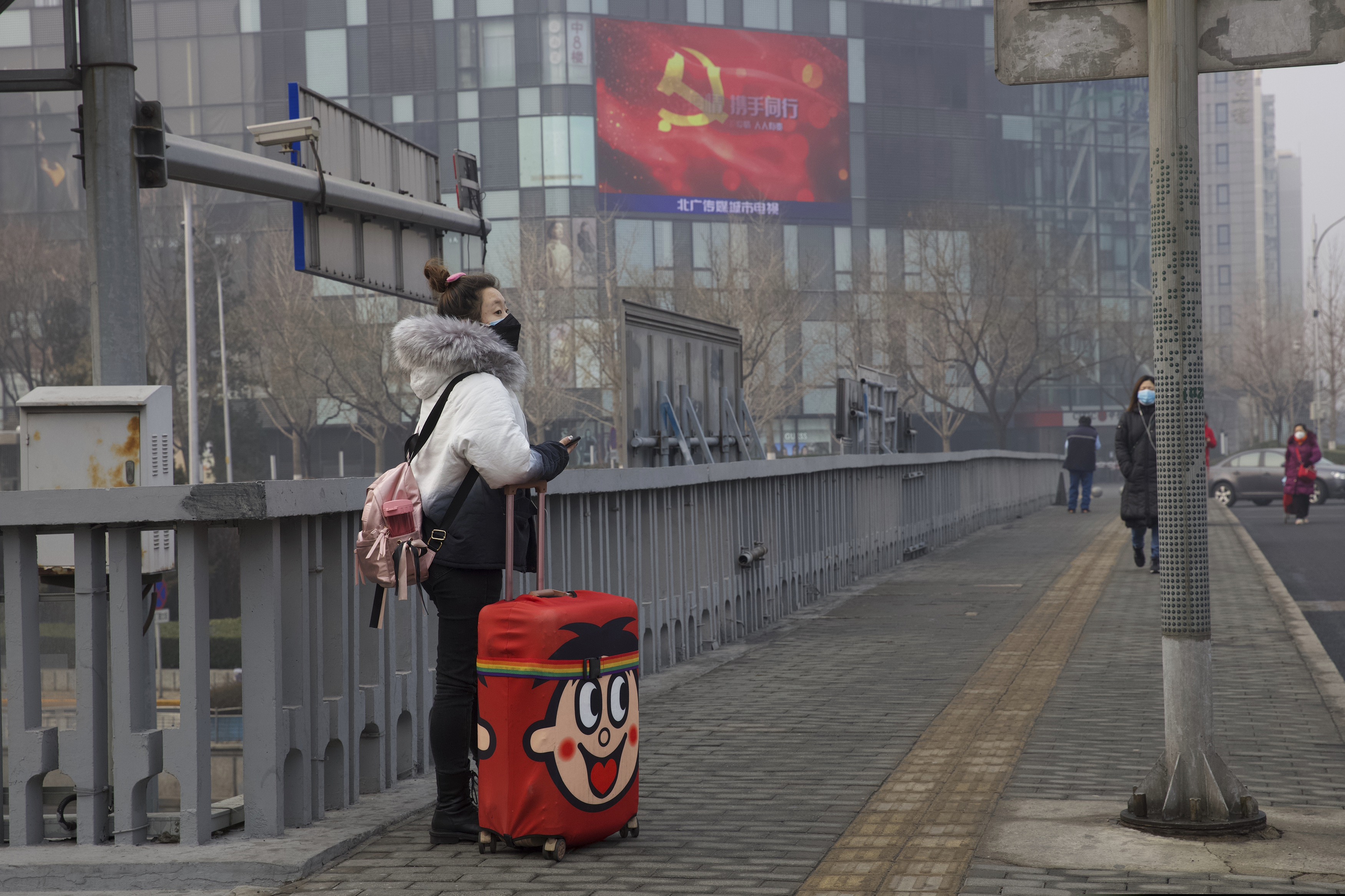 A traveler stands on a bridge near a display showing government propaganda in the fight against the COVID-19 viral illness in Beijing, China Thursday, Feb. 13, 2020. China is struggling to restart its economy after the annual Lunar New Year holiday was extended to try to keep people home and contain novel coronavirus. Traffic remained light in Beijing, and many people were still working at home.