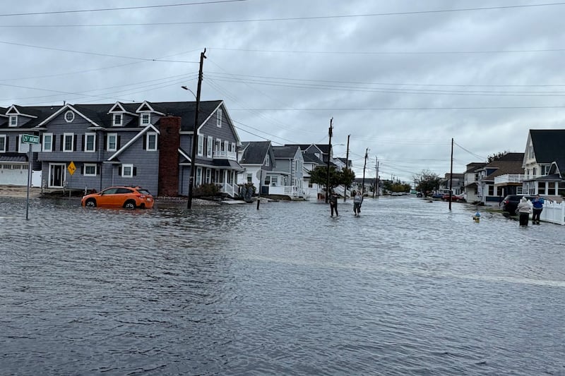 People wade through a flooded residential intersection in Wildwood, New Jersey, with water surrounding parked cars beneath cloudy skies.
