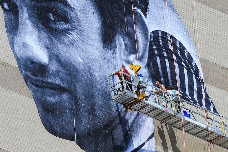 Artist JR, with fedora, installs his mural of Ibrahim, a Pakistani immigrant and Philadelphia resident, on the side of the Graham Building at 30 S. 15th St. The portrait is part of Mural Arts' "Open Source" series.