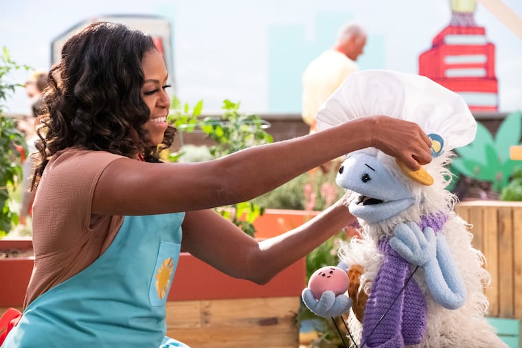 Michelle Obama puts a chef's hat on Waffles, a furry puppet with waffle ears and holding Mochi, a pink round puppet, on the set of the children's series "Waffles + Mochi."