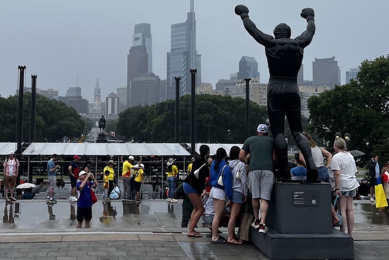 Tourists pose with the Rocky statue on the steps of the Philadelphia Museum of Art on Saturday, June 27, 2026. In the background, the concert stage for the city's July 4 concert can be seen being built by crews ahead of the show.