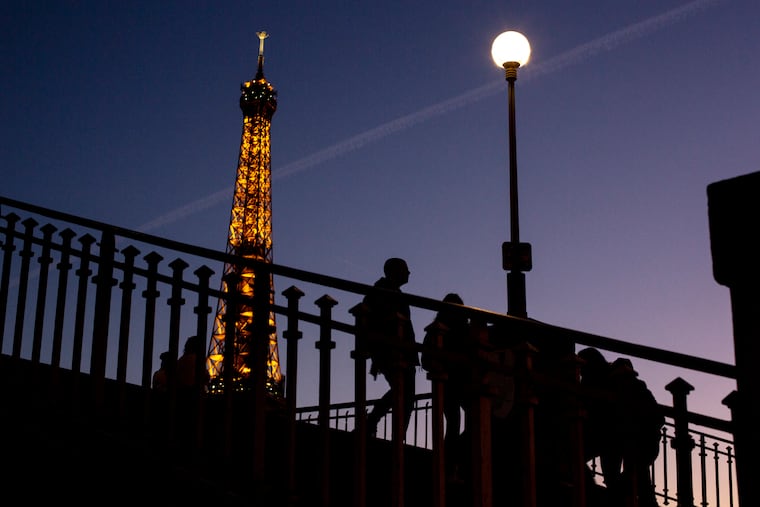 People walk on a bridge next to the Eiffel Tower in Paris, Wednesday Feb. 9, 2022. Lights on the Eiffel Tower will soon be turned off an hour earlier at night as part of an energy savings plan in the French capital, its mayor announced. Paris mayor said the iconic tower that is illuminated until 1:00am is only one of the city's monuments and municipal buildings that will be plunged into darkness earlier in the evening as the French capital faces risks of power shortages, rationing and blackouts when energy demand surges this winter.