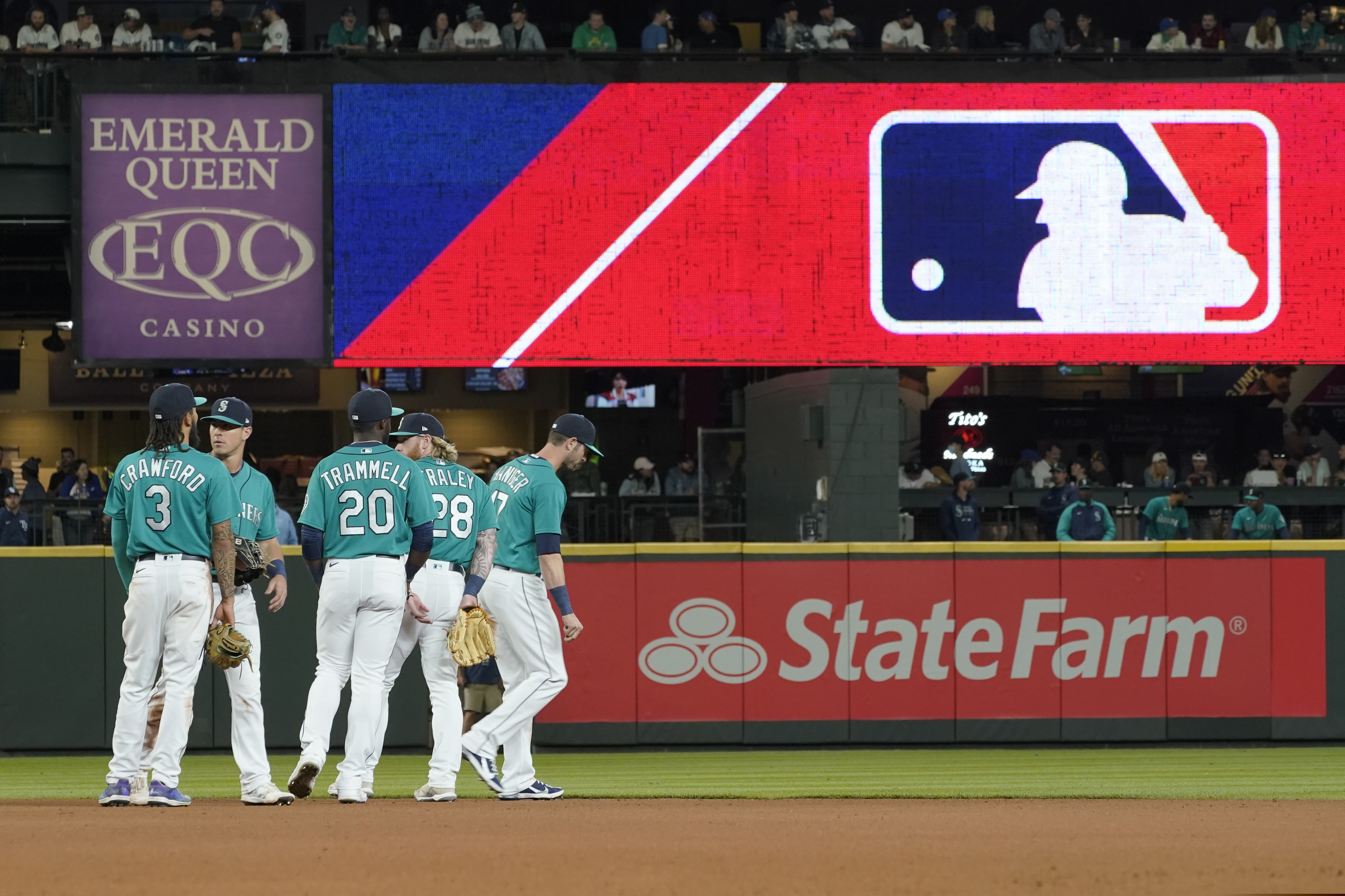 Seattle Mariners gather as the MLB logo is shown during a review of an attempted catch by right fielder Mitch Haniger of a ball hit by Tampa Bay Rays' Ji-Man Choi that was originally called an out during the ninth inning of a baseball game Friday, June 18, 2021, in Seattle. The call was overturned. While Major League Baseball and the players union try to chart a path forward, hoping to get baseball back on the field, some experts are watching the talks more from an academic viewpoint.