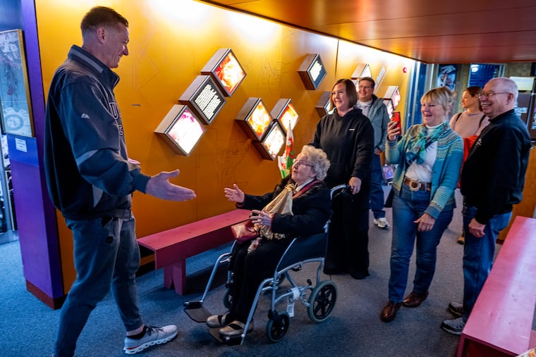 ”Queen Mumm” Avril Davidge, a 93 year old Welsh grandma is surprised by Quaker City String Band Captain Jimmy Good (left) at the Mummers Museum Wednesday. Davidge gets to live her dream of going to the Mummers Parade, starting on New Year’s Eve morning with a tour of the museum with daughter daughter Kay Hedges (right, with phone/camera) and granddaughter Fiona Smillie-Hedges (pushing wheelchair).