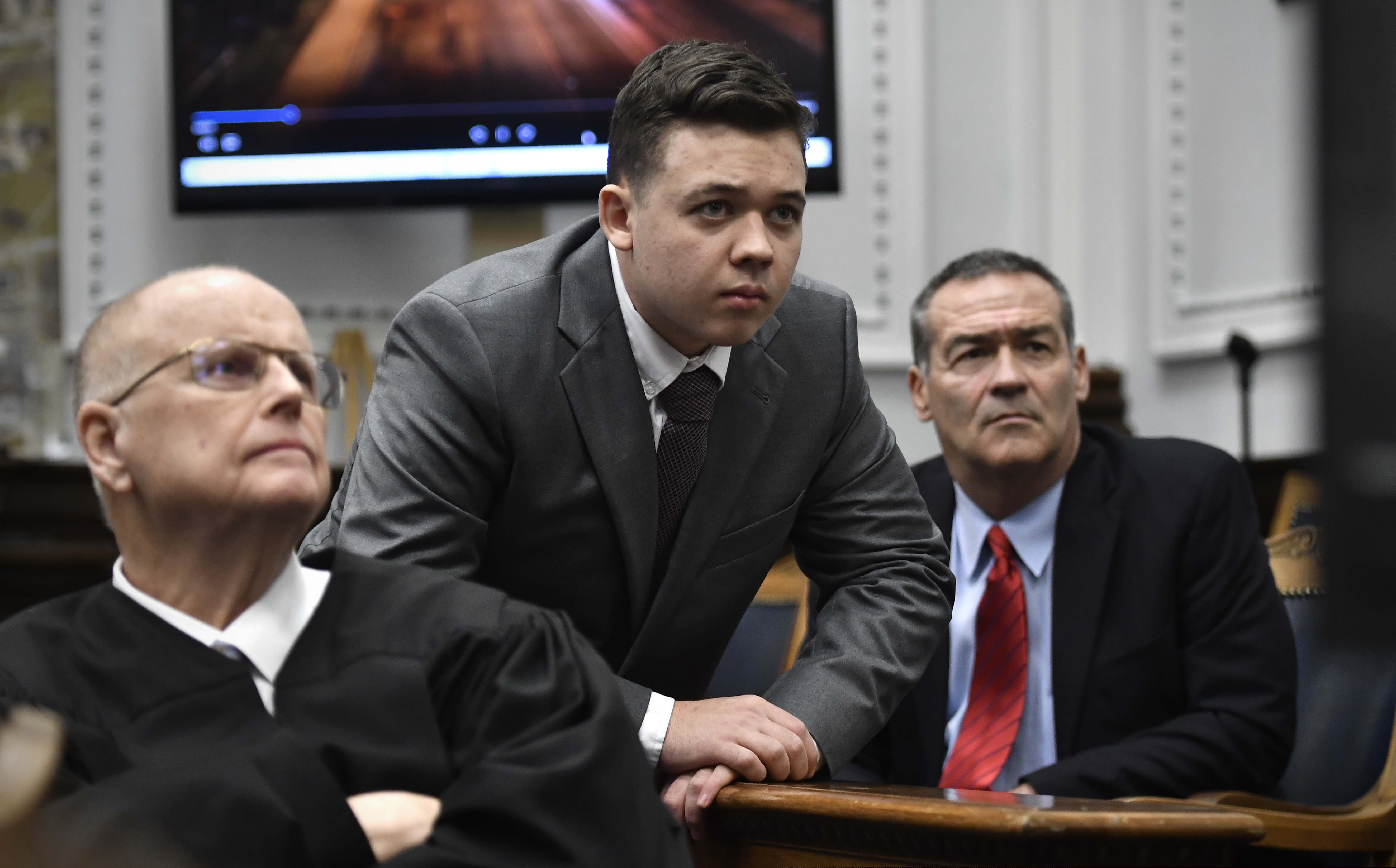 Judge Bruce Schroeder (left), Kyle Rittenhouse (center) and his attorney Mark Richards watch an evidence video in question on a screen during proceedings at the Kenosha County Courthouse in Kenosha, Wis., on Nov. 12, 2021. As Rittenhouse’s trial has played out, moments of apparent deference to the defendant by the judge have struck observers as curiously different from other murder proceedings.