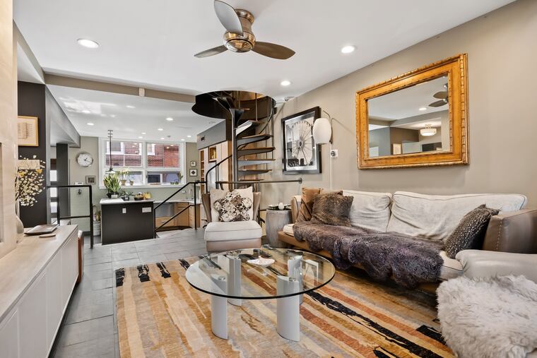 The two-story living room has 18-foot ceilings and a wall of glass overlooking a Japanese garden. It has a granite-topped bar with storage, a gas fireplace with stone surround, recessed lighting, ceiling fan, and a sliding glass door leading to the outside.