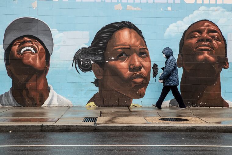 A woman wearing a surgical mask makes her way past a mural on the west side of the Save-A-Lot store at 22nd and Lehigh, where she was shopping, thats depicts many ranges of emotions on the subjects faces, on March 31, 2020.