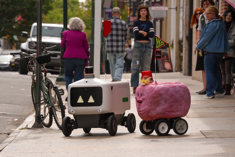 The pink, fuzzy, potato-shaped Awebot encounters an Uber Eats delivery robot in Old City, Philadelphia, Sept. 25, 2026. Awebot was created by artist Madison Polidoro.