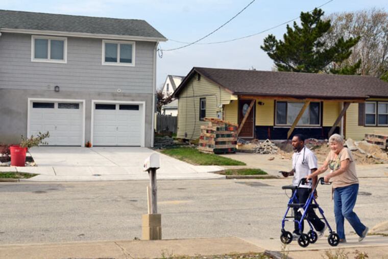Barbara Fox walks along Cummings Place in Brigantine with her physical therapist Marvin Royal on October 29, 2014, on the second anniversary of Superstorm Sandy. Her home (not shown) is similar to the house on the right. Both will be raised, just like the home on the left. ( TOM GRALISH / Staff Photographer )