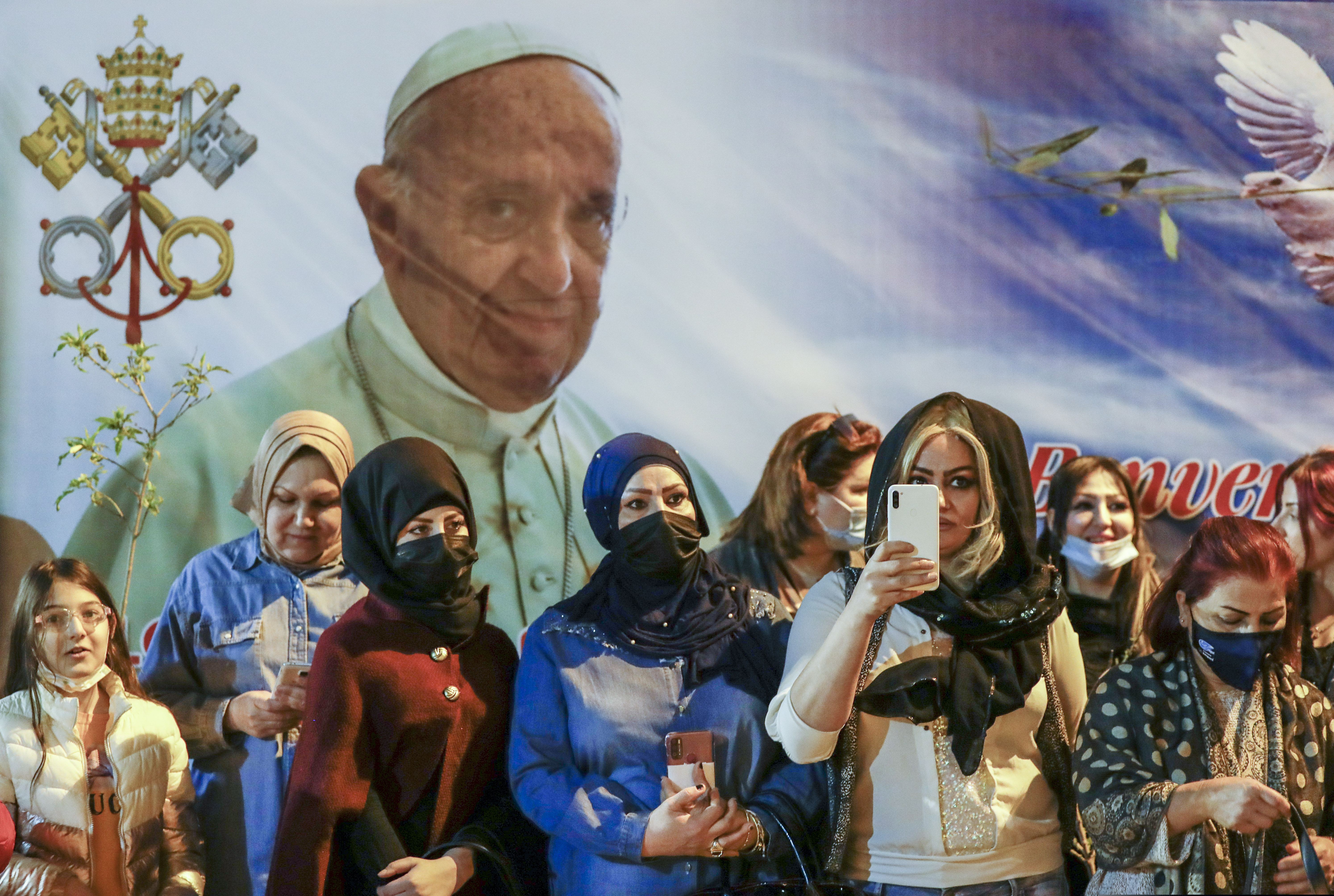 Women wait outside the Chaldean Cathedral of Saint Joseph, in Baghdad, Iraq, Saturday, March 6, 2021, where Pope Francis, depicted on a giant poster at their back, is concelebrating a mass. Earlier today Francis met privately with the country's revered Shiite leader, Grand Ayatollah Ali al-Sistani.