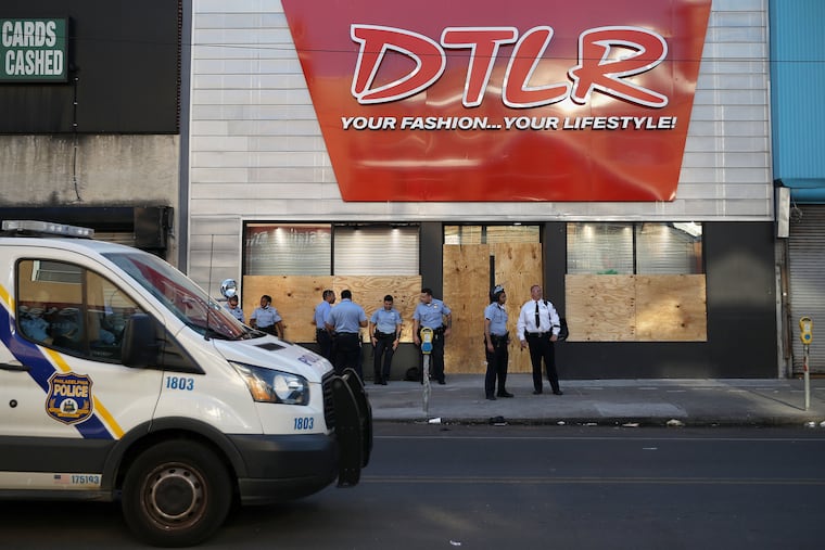 A small group of police stand outside the boarded-up DTLR store on 52nd Street near Market Street in West Philadelphia just after 6:45 p.m. on Monday, June 1, 2020. Many businesses along that stretch of 52nd Street were damaged on May 30 after property destruction and looting accompanied protests against the Minneapolis police custody death of George Floyd.