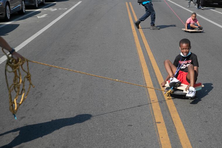Miki Baker (front) and Ny’Sir Shabazz are pushed along on “skoot boards” made for the Philadelphia Parks & Recreation Department’s annual Playstreets, after an event families and community members in Southwest Philly attended to rally against gun violence at Mitchell Elementary on Wednesday, May 19, 2021. The boards were made by the nonprofit Tiny WPA.