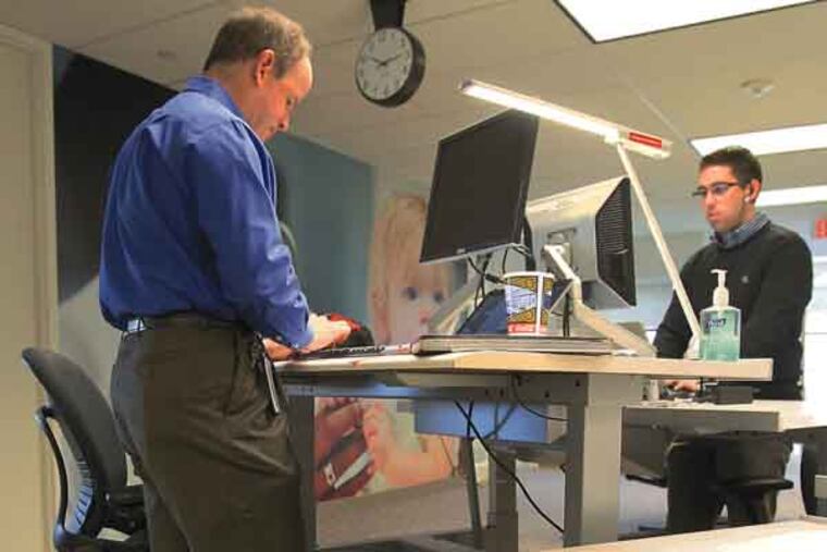 A growing body of research shows that sitting at desk jobs all day decreases life expectancy and makes you more susceptible to cancer and other diseases – and Philadelphia-area companies are taking note. And on Feb. 4, GlaxoSmithKline will move from Center City to Navy Yard, where its new office will feature height-adjustable desks, centralized printing stations and trash cans, and even treadmill work stations. The changes, the companies say, were made specifically to address health concerns and to improve productivity. Ron Joines, left, and Ken Horner of GSK work in a standing position at a height adjustable desks. ( Charles Fox / Staff Photographer )