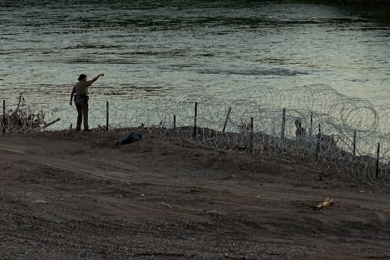 A Texas state trooper talks with migrants as they walk along concertina wire on the banks of the Rio Grande as they try to enter the U.S. from Mexico in Eagle Pass, Texas, Thursday, July 6, 2023. Texas Republican Gov. Greg Abbott has escalated measures to keep migrants from entering the U.S.