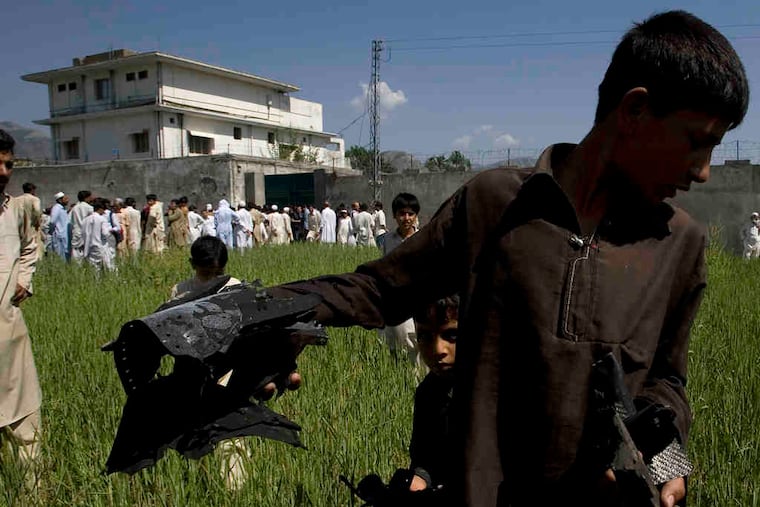 A Pakistani youth shows metal pieces , believed to be parts of the U.S. helicopter lost in the raid on Osama bin Laden's compound (background). Residents on Tuesday provided details on who lived in the compound. Story on A12.