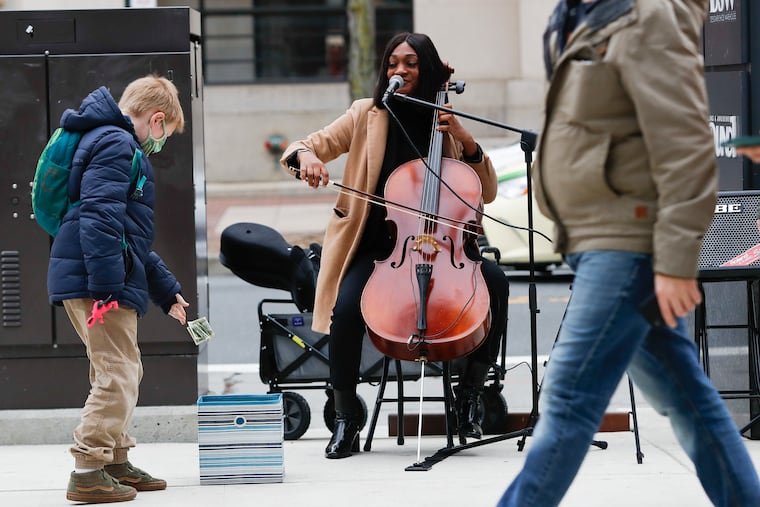 Cellist/vocalist Aijee Evans, watches Ben Maher, 9, drop dollar bills into a tip box, while Evans performed at Ninth and Market Streets in Center City on Monday, November 22, 2021. Evans was robbed in November while performing at Fourth and South Streets by a couple of teens.