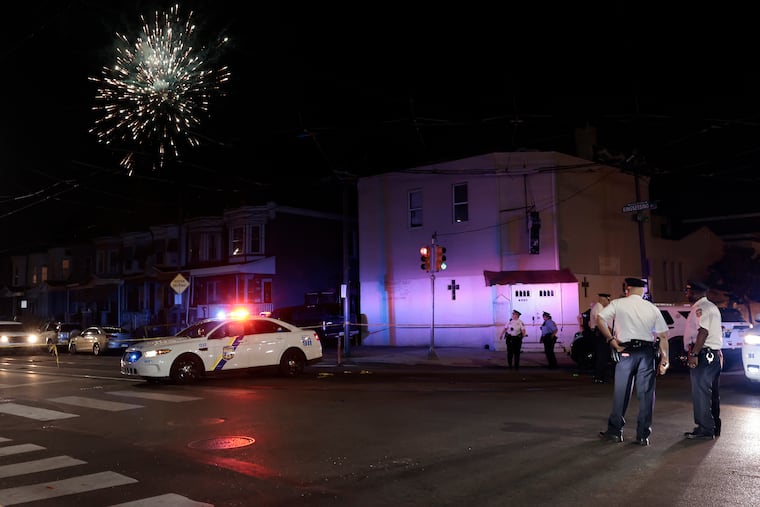 Neighborhood fireworks explode in the background as police investigate a July 4, 2024, shooting near South 60th Street and Kingsessing Avenue in Southwest Philadelphia that killed one person and injured eight others.