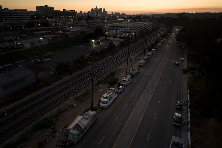 A line of RVs where unhoused people live stretches along a street as the sun sets behind the Los Angeles skyline.