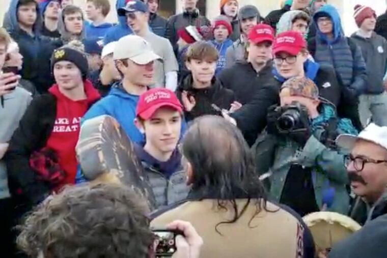 In this Friday, Jan. 18, 2019 image made from video provided by the Survival Media Agency, Nicholas Sandmann, wearing a "Make America Great Again" hat, center left, stands in front of an elderly Native American singing and playing a drum in Washington.