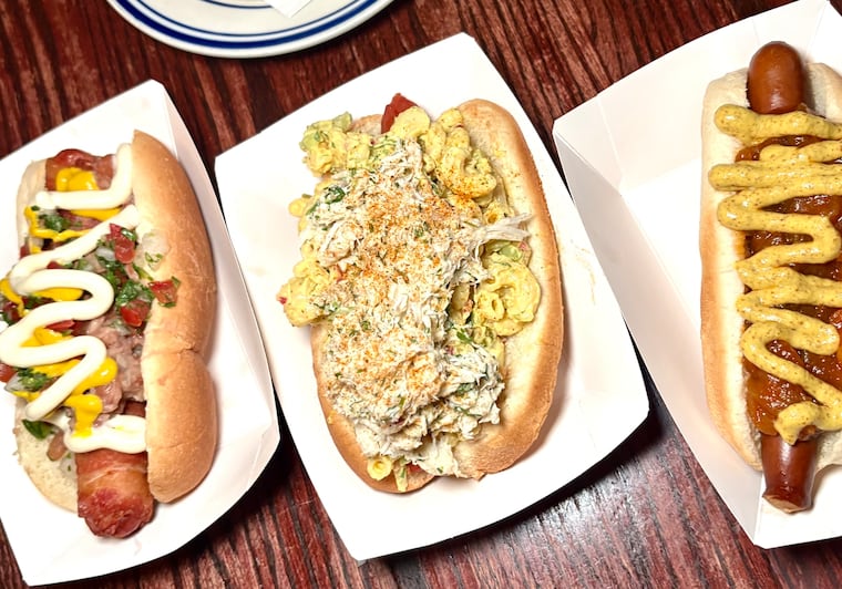 Three weekly special from Royal Tavern's July-only hot dog menu. From left to right: a fried Diamondback Dog wrapped in bacon with salsa and beans, an Old Bay Bomber topped with Old Bay macaroni salad and crab, and Barrel It Up The Bronx topped with onion sauce and brown mustard.
