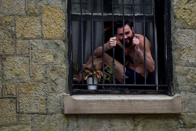 Justin Donahue mimes being in a prison cell as he says goodby to a friend who had just dropped some provisions off for him on his doorstep while he self-isolates at home on the 2200 Block of St. James Place March 22, 2010. His brother has tested positive for the coronavirus and Donahue is in Day Five of his quarantine while awaiting results of his own testing. He says he sits in the window because he is bored with being alone.