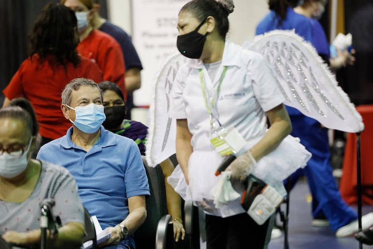 Dressed as an angel, Marisol Rivera of Camden shows her wings to Romulo Gaitan of Mount Laurel while constantly sanitizing the waiting area at the Camden County Vaccination Center at Camden County College in Blackwood , NJ on March 11, 2021. When Marisol Rivera was a waitress at the Cherry Hill Diner she would occasionally dress up in costumes to entertain her customers-until COVID-19. When some co-workers became ill she said “ I need to live-I’ve got grandkids” and quit her job as a waitress. Now with her new “I love it here” job, with Pritchard Industries, she helps to keep the Camden County Vaccination Center at Camden County College spotless while continuing her craft of making people smile and feel at ease after they get their COVID-19 vaccination.