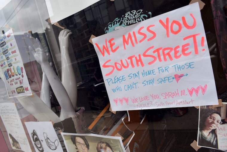 Bright neon text on a white paper hangs in the window of South Street Art Mart displaying the text: "WE MISS YOU SOUTH STREET, PLEASE STAY HOME FOR THOSE WHO CAN'T. STAY SAFE."