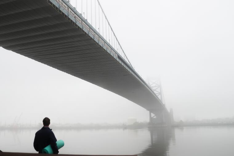A man with a yoga mat stands underneath a foggy Ben Franklin Bridge at Race Street Pier in Philadelphia on Tuesday, Oct. 20, 2020.