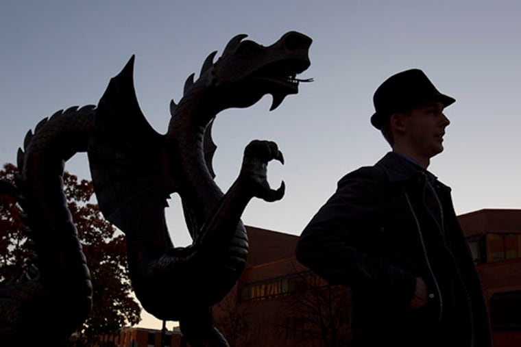 Drexel senior Arsen Nikiforouk is silhouetted as he walks by the statue of Mario the Magnificent, the Drexel Dragon, at 33rd and Market Streets as the sun sets. ( CHARLES FOX / Staff Photographer )
