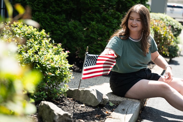 Kelly Harrington, 29, touches an American flag on a neighbor’s lawn while posing for a portrait near her home in Conshohocken, Pa., on Monday, Aug. 24, 2026. She recently participated in an Inquirer/NYT/Siena poll.